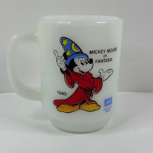 May include: White ceramic mug featuring Mickey Mouse in a sorcerer's outfit from Fantasia. The design includes Mickey Mouse in a red robe and blue hat with stars. Text on the mug reads "Mickey Mouse in Fantasia" and "1940".
