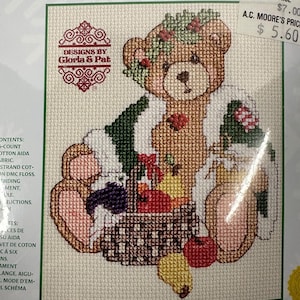 Peut inclure: Kit de point de croix représentant un ours en peluche brun avec une couronne florale, tenant un panier de fruits. Le motif utilise des couleurs vertes, rouges et violettes sur une toile Aida crème. Le kit est de Designs by Gloria & Pat.