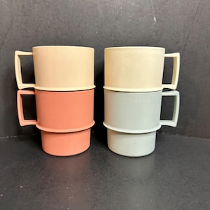 May include: Two stacks of two mugs each, featuring a matte finish. The left stack includes a cream-colored mug on top of a terracotta-colored mug. The right stack features a cream-colored mug on top of a sage green mug. Each mug has a square handle.