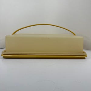 Puede incluir: Una mantequera rectangular con una tapa de color crema claro y una base de color amarillo mostaza. El recipiente incluye un asa arqueada a juego de color amarillo mostaza que se extiende sobre la tapa, proporcionando un agarre funcional.