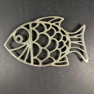 Può includere: Un sottopentola in metallo dorato a forma di pesce, caratterizzato da un design traforato con linee curve che formano la testa, il corpo e le pinne caudali. La superficie ha una finitura lucida e riflettente, composta da anelli metallici.