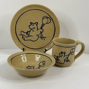 May include: A tan ceramic dish set featuring a plate, bowl, and mug. Each piece has a dark blue outline drawing of a bear holding balloons. The plate and bowl have a dark blue border. The mug has a handle.