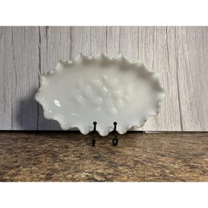Puede incluir: Un plato blanco ovalado con borde ondulado y diseño floral. El plato es de vidrio opaco y se exhibe sobre un soporte de metal negro. El fondo presenta una pared de madera.