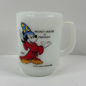 May include: White ceramic mug featuring Mickey Mouse in a red robe and blue wizard hat from the movie Fantasia. The mug has a handle and the text "MICKEY MOUSE in FANTASIA" and "1940". The mug is a vintage collectible.