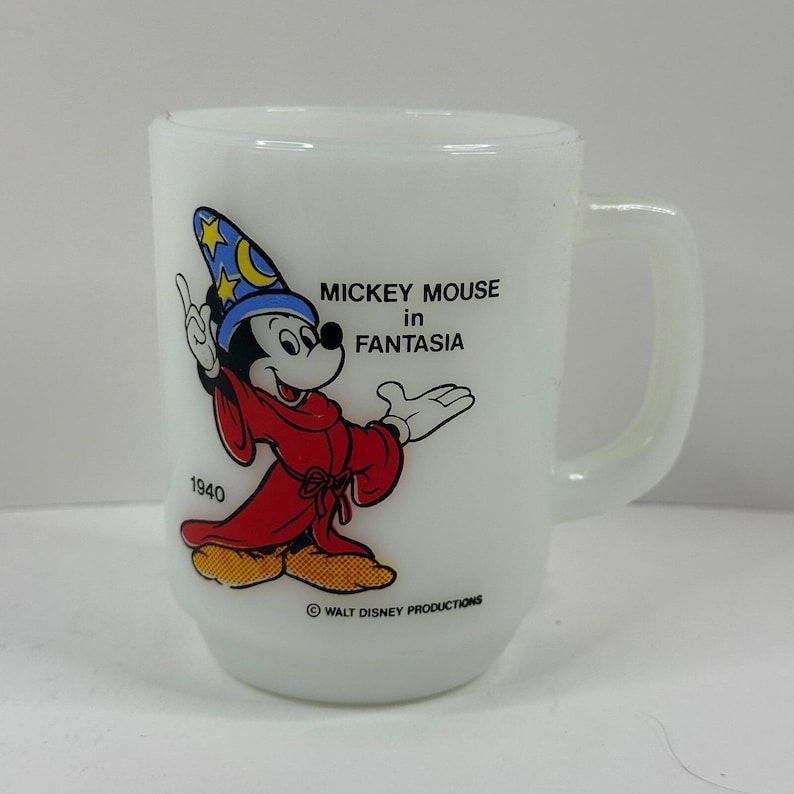 May include: White ceramic mug featuring Mickey Mouse in a red robe and blue wizard hat from the movie Fantasia. The mug has a handle and the text "MICKEY MOUSE in FANTASIA" and "1940". The mug is a vintage collectible.