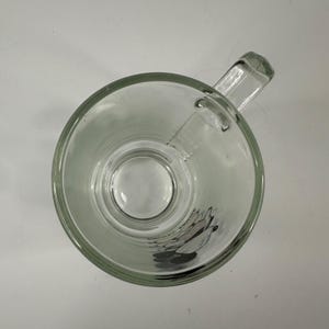 May include: A clear glass mug viewed from directly above, featuring a sturdy handle on the right side. The bottom interior of the mug displays a graphic of Mickey Mouse with the text Mickey Mouse printed in black lettering near the character.