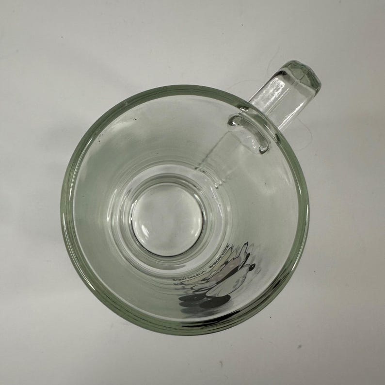 May include: A clear glass mug viewed from directly above, featuring a sturdy handle on the right side. The bottom interior of the mug displays a graphic of Mickey Mouse with the text Mickey Mouse printed in black lettering near the character.