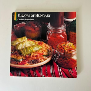 May include: A cookbook titled "Flavors of Hungary" by Charlotte Slovak Biro. The cover features a wooden bowl of stuffed cabbage, jars of pickled vegetables, and a red and white patterned tablecloth.