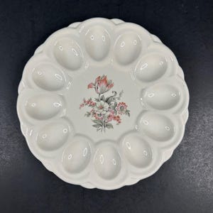 May include: White ceramic egg serving platter with 12 egg-shaped indentations. The center features a floral design with pink, red, and gray flowers. The platter has a scalloped edge.