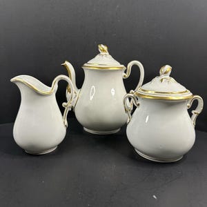 May include: A three-piece porcelain tea set. The set includes a teapot with a lid, a creamer, and a sugar bowl with a lid. Each piece is white with gold trim and accents.