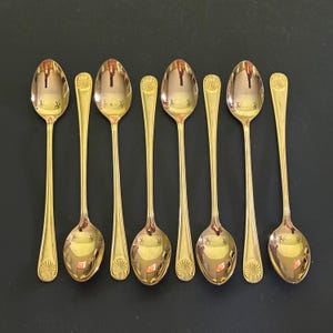 Puede incluir: Ocho cucharaditas doradas con un diseño decorativo de concha en el mango. Las cucharitas están dispuestas en una fila sobre una superficie oscura.