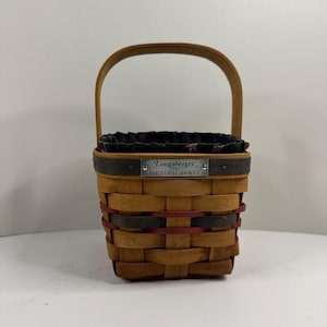 Pode incluir: Uma cesta tecida com uma alça de madeira e forro de tecido preto. A cesta tem formato retangular com uma etiqueta de metal que diz "Longaberger 1993 Inaugural Basket". A cesta é feita de material tecido marrom claro e marrom escuro.