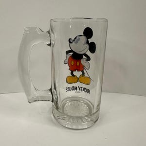 Peut inclure: Une chope &agrave; bi&egrave;re en verre transparent orn&eacute;e d'une illustration de Mickey Mouse portant un short rouge et des chaussures jaunes. Le personnage se tient les mains sur les hanches. Sous l'image, le texte MICKEY MOUSE est imprim&eacute; en lettres capitales noires.