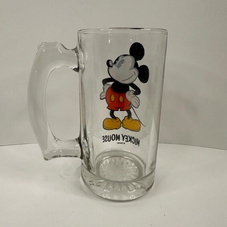 Peut inclure: Une chope &agrave; bi&egrave;re en verre transparent orn&eacute;e d'une illustration de Mickey Mouse portant un short rouge et des chaussures jaunes. Le personnage se tient les mains sur les hanches. Sous l'image, le texte MICKEY MOUSE est imprim&eacute; en lettres capitales noires.