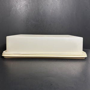 Puede incluir: Un recipiente de almacenamiento rectangular de plástico color blanco hueso con una tapa a juego. El recipiente presenta un acabado liso y opaco, con un diseño sencillo y funcional que incluye un borde ligeramente elevado en la base.