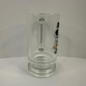 May include: A clear glass mug featuring a side profile graphic of Mickey Mouse with black ears, a white face, and yellow shoes. The text MICKEY MOUSE is printed in black below the character. The mug has a sturdy handle and a thick, reinforced base.