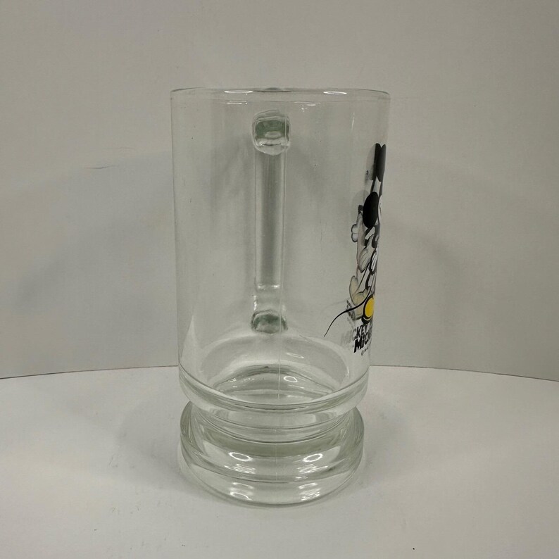 May include: A clear glass mug featuring a side profile graphic of Mickey Mouse with black ears, a white face, and yellow shoes. The text MICKEY MOUSE is printed in black below the character. The mug has a sturdy handle and a thick, reinforced base.