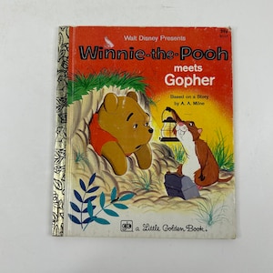 Könnte beinhalten: Ein Vintage-Kinderbuchcover mit Winnie Puuh und Gopher. Das Cover ist rot und gelb mit dem Titel "Winnie Puuh trifft Gopher" in großen, fetten Buchstaben. Die Illustration zeigt Winnie Puuh, der aus einem Loch in einem Baum lugt, während Gopher eine Laterne und eine Werkzeugkiste hält.