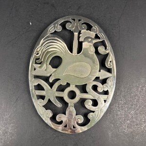 Puede incluir: Un salvamanteles ovalado de metal plateado con un diseño recortado de un gallo de pie sobre una flecha. El marco decorativo incluye las letras N y S de los puntos cardinales, junto con patrones de volutas ornamentales alrededor de la figura.