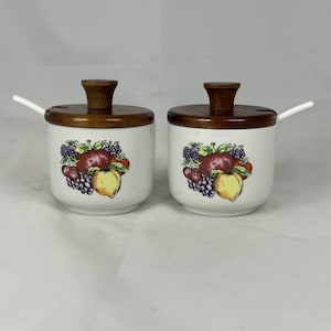 May include: Two white ceramic condiment jars with wooden lids and small white spoons. Each jar features a colorful fruit design with apples, grapes, and peaches. The lids are a dark brown color.