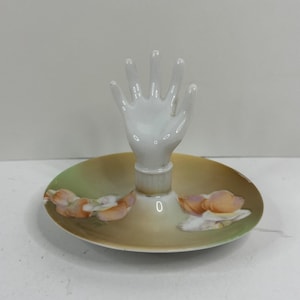 May include: A white porcelain hand-shaped ring holder with a round, floral-decorated base. The base features a gradient of green and yellow with painted peach-colored flowers. The ring holder is a decorative item.