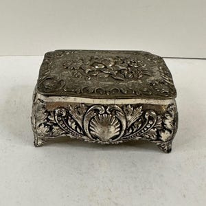 Peut inclure: Une boîte à bijoux rectangulaire en métal argenté avec un motif floral en relief sur le couvercle et un motif de coquillage sur le panneau avant. La boîte repose sur quatre petits pieds et présente des sculptures complexes en forme de feuilles.