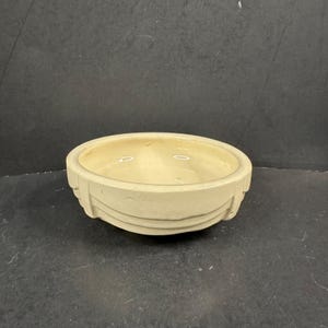 Puede incluir: Un cuenco redondo de cerámica de color crema con un acabado interior liso y brillante. El exterior presenta una serie de bandas horizontales en relieve que envuelven la circunferencia, creando un sutil elemento de diseño arquitectónico.