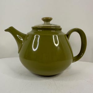 May include: Olive green ceramic teapot with a rounded body, curved handle, and a spout. The lid is a lighter shade of green with a small knob. The teapot is designed for brewing tea and has a glossy finish.