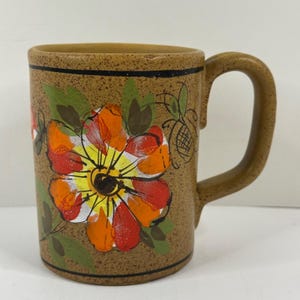 May include: A brown ceramic mug with a large, hand-painted floral design in shades of red, orange, and yellow. The mug has a speckled texture and a curved handle. Green leaves and a black border complete the design.