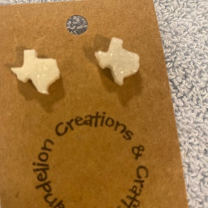 May include: A pair of white glitter Texas-shaped earrings on a brown card with the text "Dandelion Creations & Crafts".