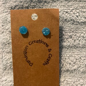 May include: A pair of blue glitter stud earrings on a brown card with the text "Dandelion Creations & Crafts".