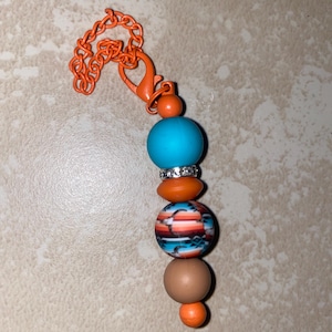 May include: A beaded keychain with an orange chain, a turquoise bead, a small orange bead with rhinestones, a patterned bead with orange, blue, and white stripes, a brown bead, and a small orange bead.
