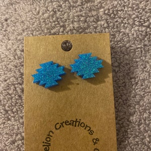 May include: A pair of blue glitter earrings with a geometric design. The earrings are on a brown card with the text "Dandelion Creations & Crafts".