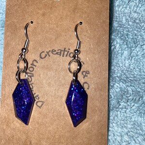 May include: A pair of dangle earrings with a purple glitter resin teardrop shape. The earrings have silver hooks.