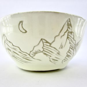Op de afbeelding: Crèmekleurige keramische kom met een handgetekend bergketen- en maanontwerp. De kom heeft een licht uitlopende rand en een gladde, glanzende afwerking. Het bergontwerp is in grijstinten.