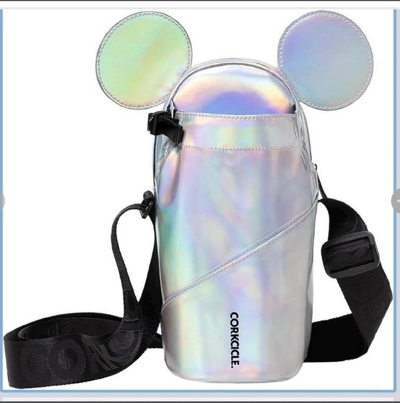 Może przedstawiać: Iridescentna srebrna torba na ramię z dwoma okrągłymi, uszowatymi akcentami. Torba ma czarny, regulowany pasek i czarne zapięcie na klamrę. Nazwa marki, CORKCICLE., jest wydrukowana na boku torby w kolorze czarnym.