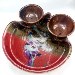 May include: A handcrafted ceramic serving dish with three attached bowls. The dish features a vibrant color palette of red, brown, blue, and green. The serving dish is ideal for serving appetizers or snacks.