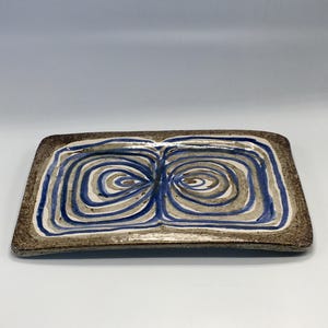 May include: Rectangular ceramic serving dish with a brown border and a central design of blue, white, and tan concentric swirls. The dish has a slightly raised edge and a matte finish.