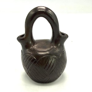 May include: A dark brown ceramic vessel with a handle and two spouts. The body of the vessel features a woven pattern design. The handle arches over the top, creating a loop. The vessel is likely used for holding liquids.