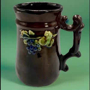 May include: A brown ceramic mug with a decorative handle. The mug features a raised relief design of grapes and leaves in shades of blue and green. The mug has a dark brown interior and a glossy finish.