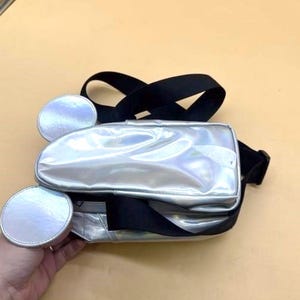 Może przedstawiać: Srebrna nerka z czarnymi paskami i dwoma okrągłymi, srebrnymi akcentami. Torba ma błyszczące, metaliczne wykończenie i prostokątną komorę gł&oacute;wną. Paski są regulowane.