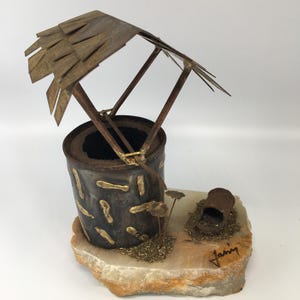 May include: A rustic metal well sculpture with a weathered appearance. The well features a small roof, a cylindrical body, and decorative accents. It rests on a stone base with a small, decorative element. The sculpture has a vintage aesthetic.