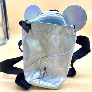 Może przedstawiać: Srebrny, opalizujący plecak z czarnymi paskami i detalami uszu myszy. Torba ma małą kieszeń z przodu, a nazwa marki "Corkcicle" jest nadrukowana z boku. Torba wykonana jest z błyszczącego, odblaskowego materiału.