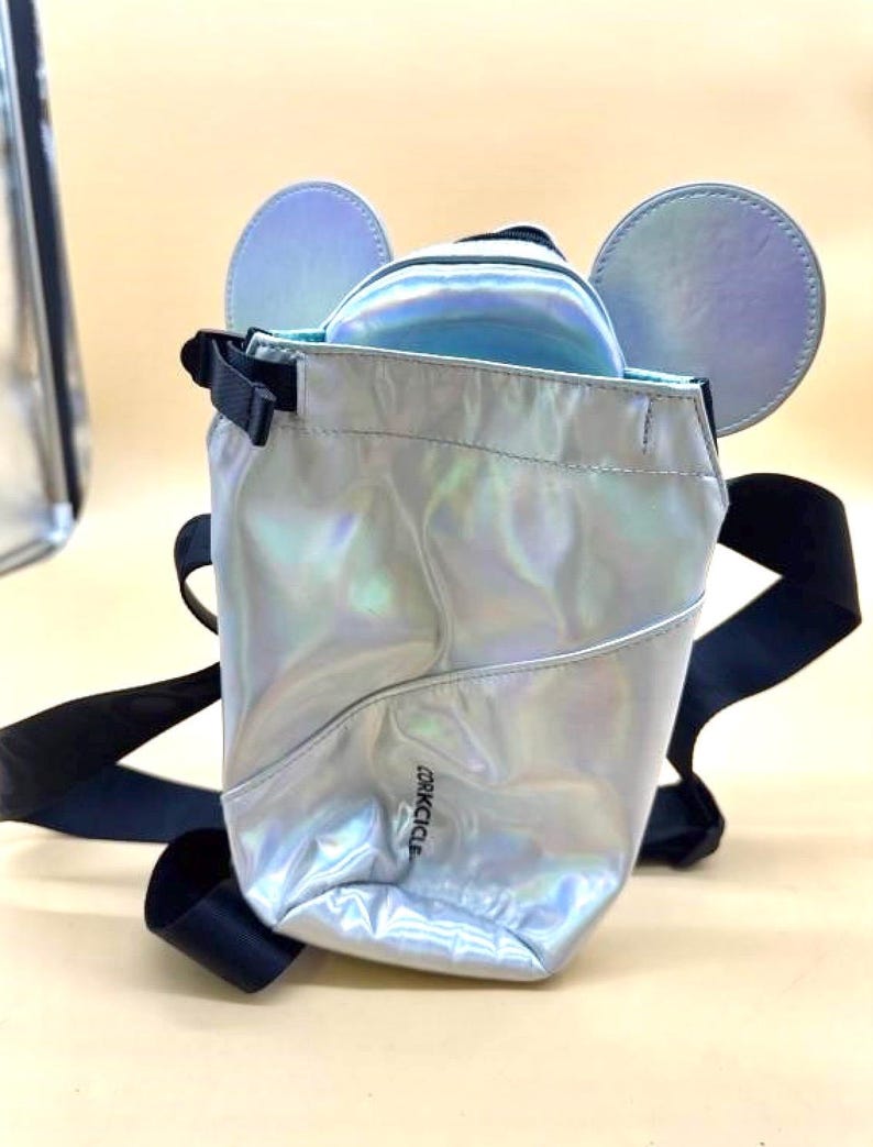 Może przedstawiać: Srebrny, opalizujący plecak z czarnymi paskami i detalami uszu myszy. Torba ma małą kieszeń z przodu, a nazwa marki "Corkcicle" jest nadrukowana z boku. Torba wykonana jest z błyszczącego, odblaskowego materiału.