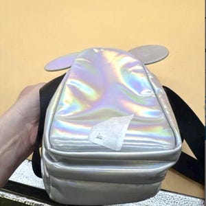Może przedstawiać: Iridescentny srebrny plecak z dołączonymi szarymi uszami myszy. Mały plecak ma czarne paski i prostokątny kształt. Powierzchnia odbija wiele kolor&oacute;w. Z przodu przymocowana jest biała naklejka.