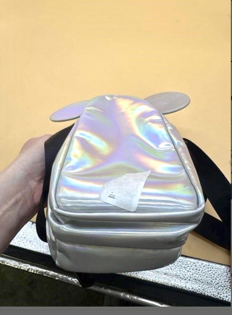 Może przedstawiać: Iridescentny srebrny plecak z dołączonymi szarymi uszami myszy. Mały plecak ma czarne paski i prostokątny kształt. Powierzchnia odbija wiele kolor&oacute;w. Z przodu przymocowana jest biała naklejka.