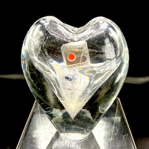 May include: A clear, heart-shaped glass sculpture with a pointed base. The sculpture has a sticker that reads "BLENKO HANDMADE" with a red circle. The glass is transparent, allowing light to pass through, and is displayed on a clear stand.