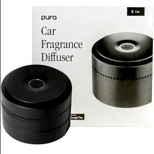 May include: Black car fragrance diffuser with the text "pura Car Fragrance Diffuser" on the packaging. The diffuser is cylindrical with a perforated band around the middle. The packaging also has a Bluetooth car icon.