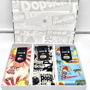 May include: A gift box containing three pairs of colorful socks. Each pair features a unique design: a popsicle, a sun with the text "Good Humor", and a tropical scene with a popsicle. The box has a popsicle-themed design.