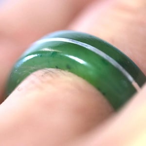 May include: A smooth, polished green jade ring featuring a thin, horizontal silver-colored inlay band running through the center of the circular stone. The ring has a rounded, domed profile and a glossy finish, showcasing the natural green color.